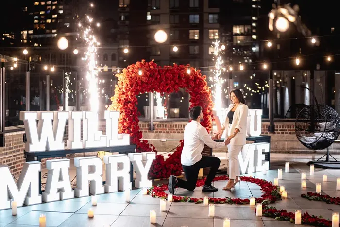 A night-time rooftop proposal in New York City — illuminated marquee letters, a red rose heart arch, cold spark fountains and candlelight