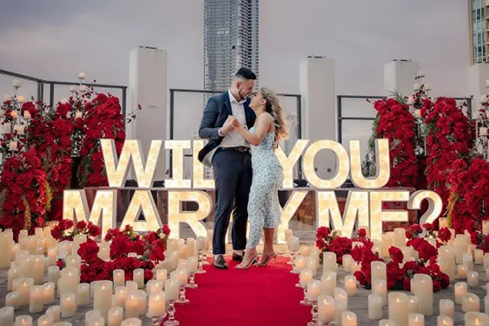 A rooftop proposal in New York City at dusk — illuminated WILL YOU MARRY ME letters, tall red rose arrangements, hundreds of candles and a couple embracing on a red runner